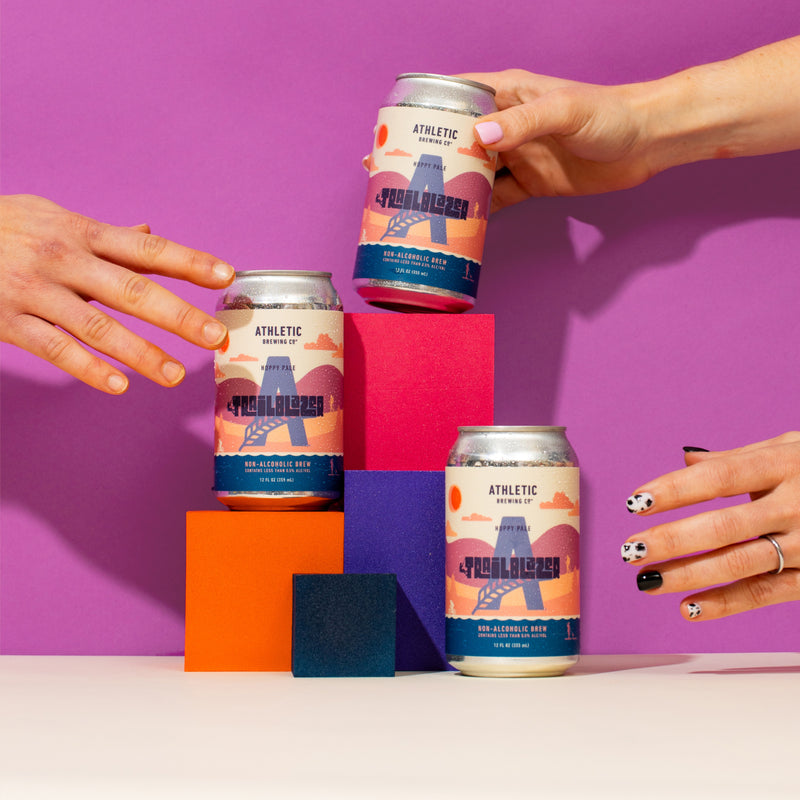 Three Trailblazer non-alcoholic beer cans arranged on colorful stacked blocks with several hands reaching in to hold or touch the cans against a purple background.