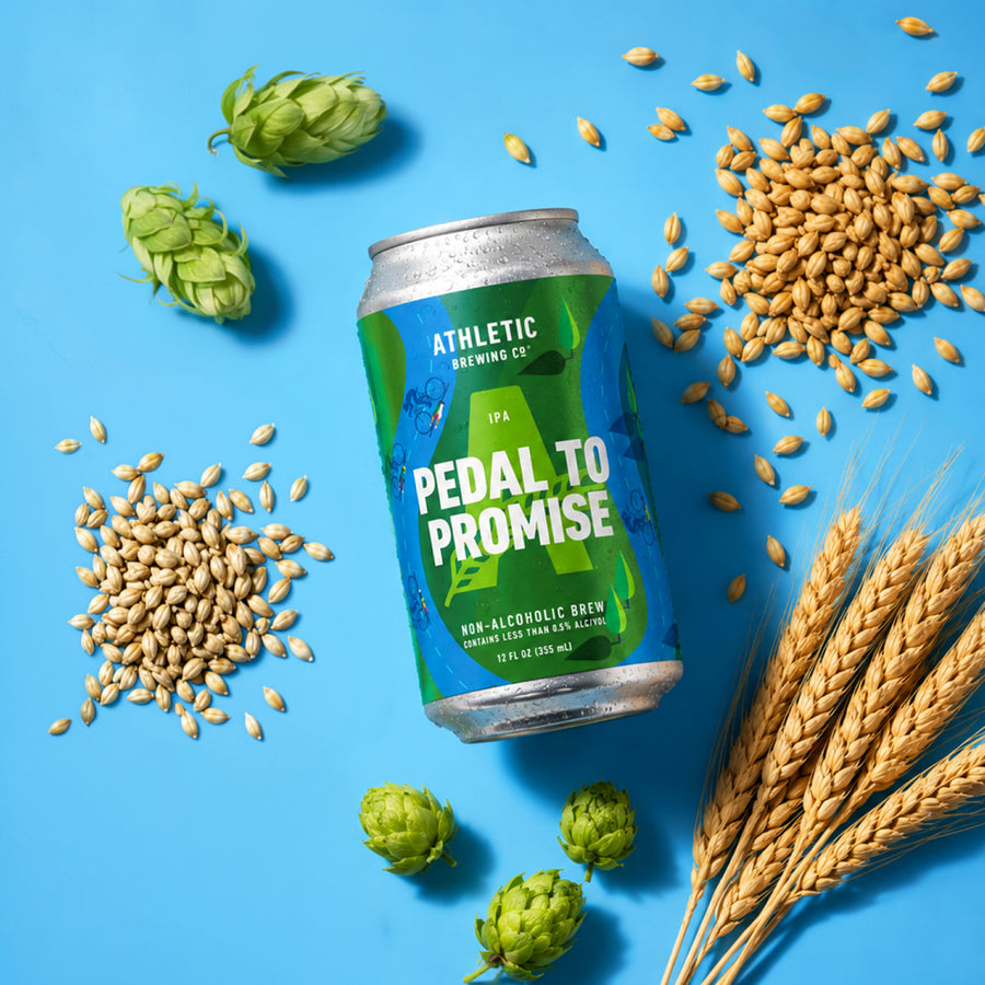 Athletic Brewing Pedal to Promise non-alcoholic IPA can lying at an angle on a bright blue surface, surrounded by green hops, scattered barley grains, and stalks of golden wheat.