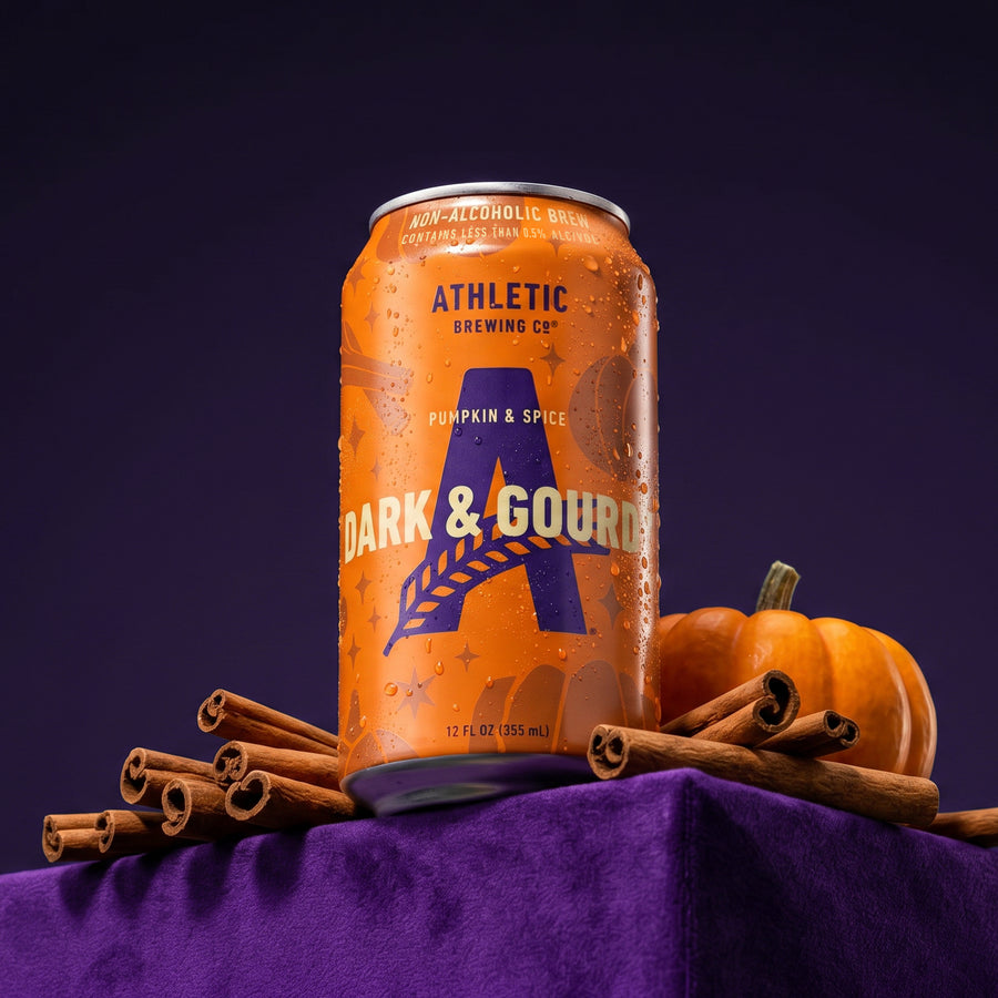Athletic Brewing Dark & Gourdy Pumpkin & Spice non-alcoholic brew can with cinnamon sticks and a pumpkin against a deep purple background.