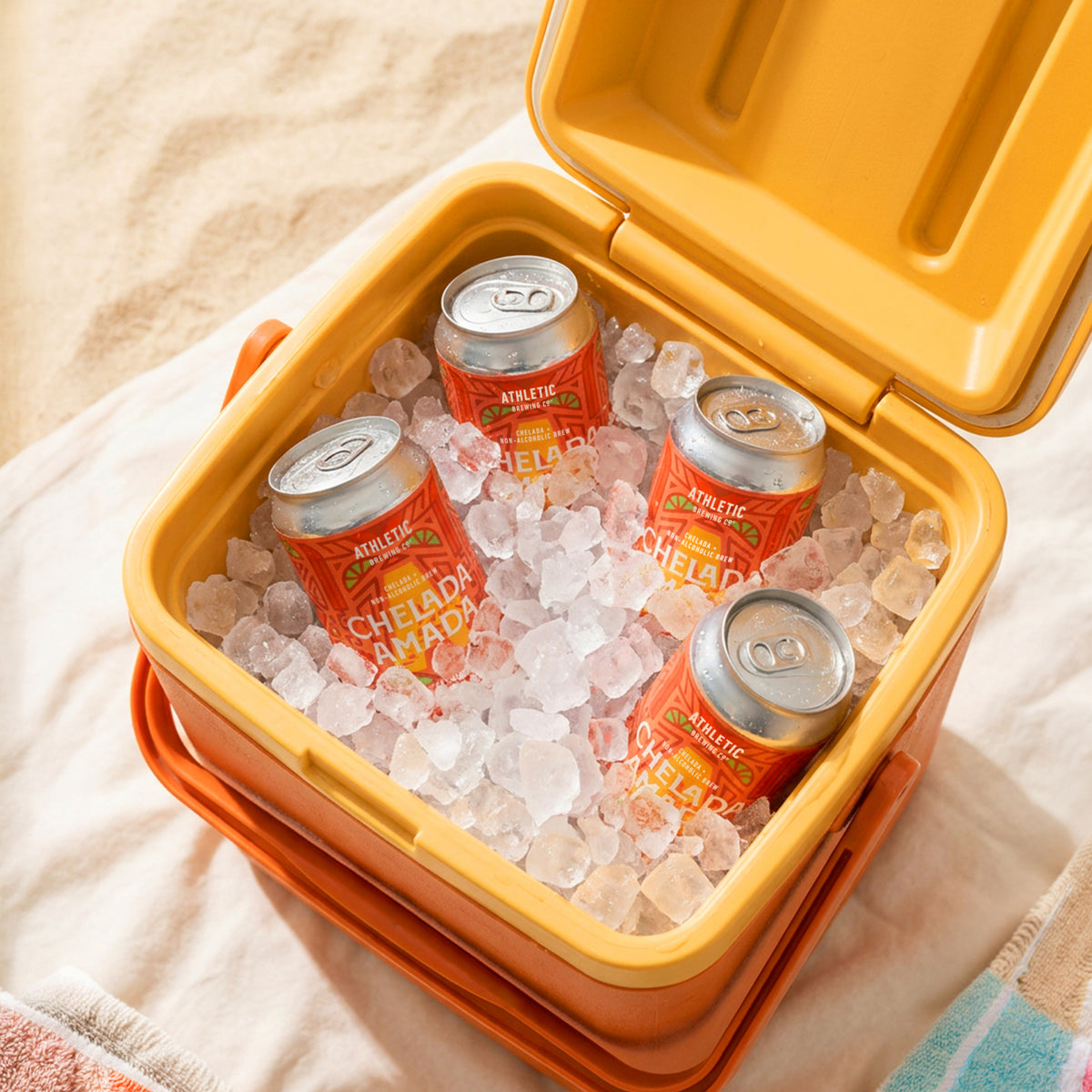 Four cans of Athletic Brewing Chelada Amada non-alcoholic brew nestled in crushed ice inside an open orange cooler. The cans are covered in condensation and arranged for easy grabbing, creating a refreshing outdoor scene on a light-colored blanket with a striped towel nearby.