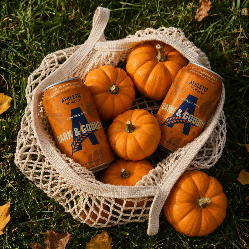 Two Athletic Brewing Dark & Gourdy non-alcoholic brew cans with small pumpkins in a market bag on grass.