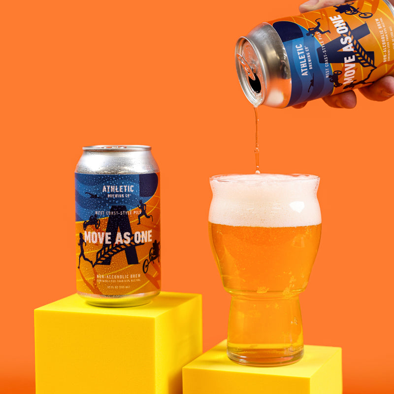 Athletic Brewing Move As One non-alcoholic pilsner being poured into a glass beside a branded can, displayed on bright yellow platforms against an orange background.