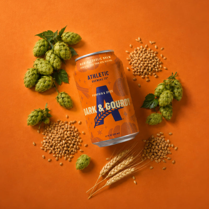 Athletic Brewing Dark & Gourdy Pumpkin & Spice non-alcoholic brew can surrounded by hops, grains, and wheat on an orange background.